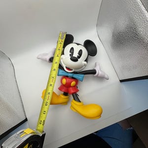 以下が含まれることがあります： 黒い頭と耳、白い顔、赤いズボンが特徴のミッキーマウスのフィギュア。 黄色の靴と青い蝶ネクタイをしています。 黄色の巻尺は、フィギュアの高さが約30cmであることを示しています。