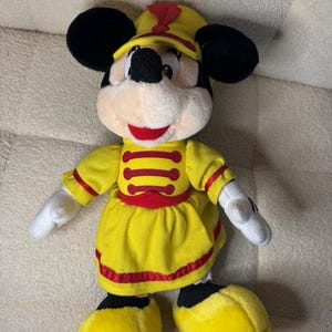 May include: A plush Minnie Mouse doll wearing a yellow marching band uniform with red accents. The doll has black ears, a black nose, and a red mouth. The uniform includes a hat, jacket, and skirt.