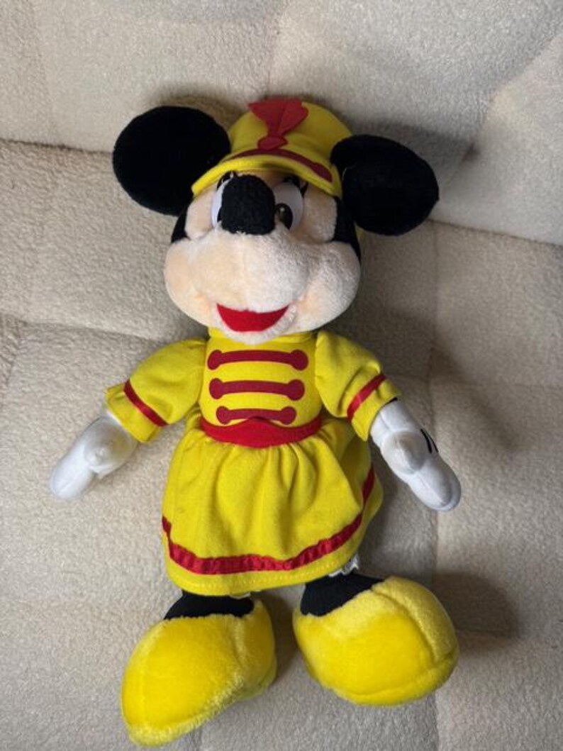 May include: A plush Minnie Mouse doll wearing a yellow marching band uniform with red accents. The doll has black ears, a black nose, and a red mouth. The uniform includes a hat, jacket, and skirt.