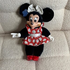 May include: A Minnie Mouse plush doll with black fur, large ears, and a white bow. She wears a red and white polka dot dress and red shoes. The doll has white gloves and is posed with one arm extended.
