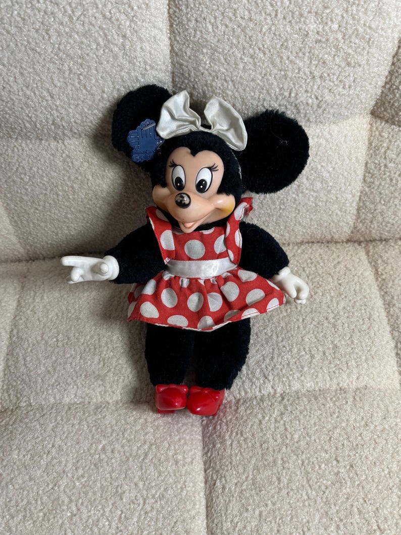 May include: A Minnie Mouse plush doll with black fur, large ears, and a white bow. She wears a red and white polka dot dress and red shoes. The doll has white gloves and is posed with one arm extended.