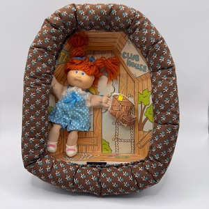 May include: A vintage doll with red hair and blue eyes, wearing a blue polka dot outfit, inside a brown and blue patterned oval-shaped bed. The doll is positioned in front of a cartoon clubhouse scene.