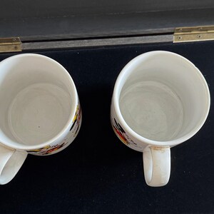 May include: Two white ceramic mugs with colorful abstract designs. The mugs have handles and are empty.