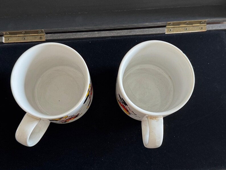 May include: Two white ceramic mugs with colorful abstract designs. The mugs have handles and are empty.