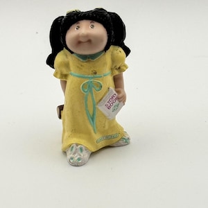 May include: A small figurine of a doll with black pigtails wearing a yellow dress with a blue ribbon. The doll is holding a small book that says "Story Book". The figurine is a collectible item.