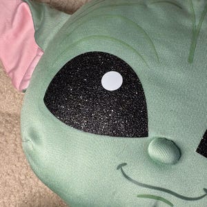 May include: A plush, green character with large black eyes and a white pupil. The character has large ears with pink interiors and a small, smiling mouth. The fabric has a textured appearance.