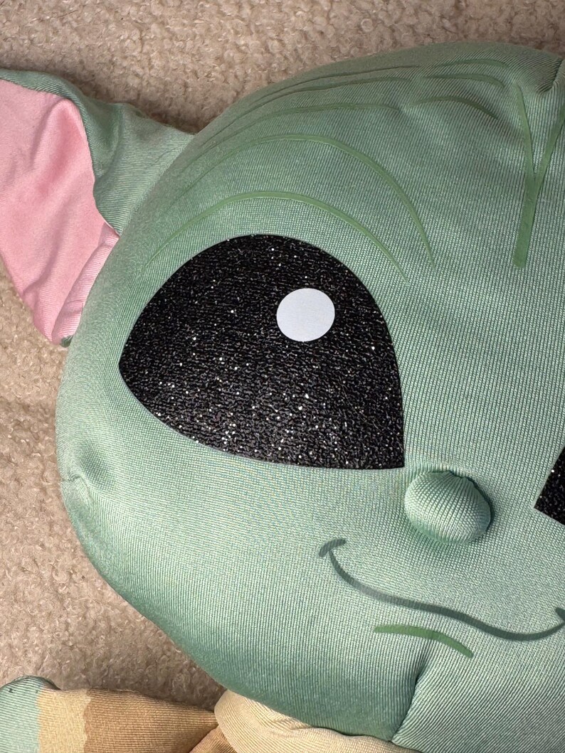 May include: A plush, green character with large black eyes and a white pupil. The character has large ears with pink interiors and a small, smiling mouth. The fabric has a textured appearance.