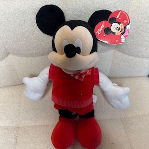 May include: A plush Mickey Mouse toy with black ears, a cream-colored face, and a red outfit. The toy has white gloves and red shoes. A heart-shaped tag with the Disney logo is attached.