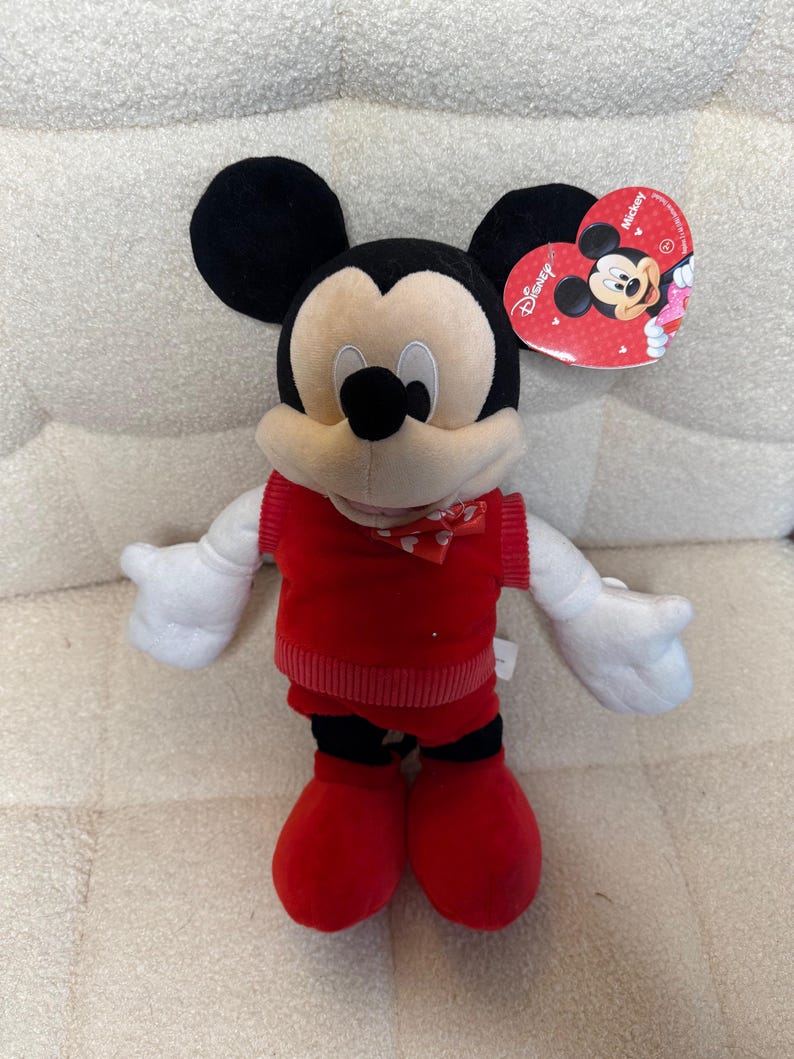 May include: A plush Mickey Mouse toy with black ears, a cream-colored face, and a red outfit. The toy has white gloves and red shoes. A heart-shaped tag with the Disney logo is attached.