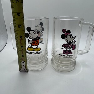 May include: Two clear glass mugs featuring Mickey Mouse and Minnie Mouse. Mickey is in his classic outfit, and Minnie is in a polka-dot dress. Each mug has a handle and the text "MICKEY MOUSE" and "MINNIE MOUSE" printed on them. The mugs are approximately 4 inches tall.