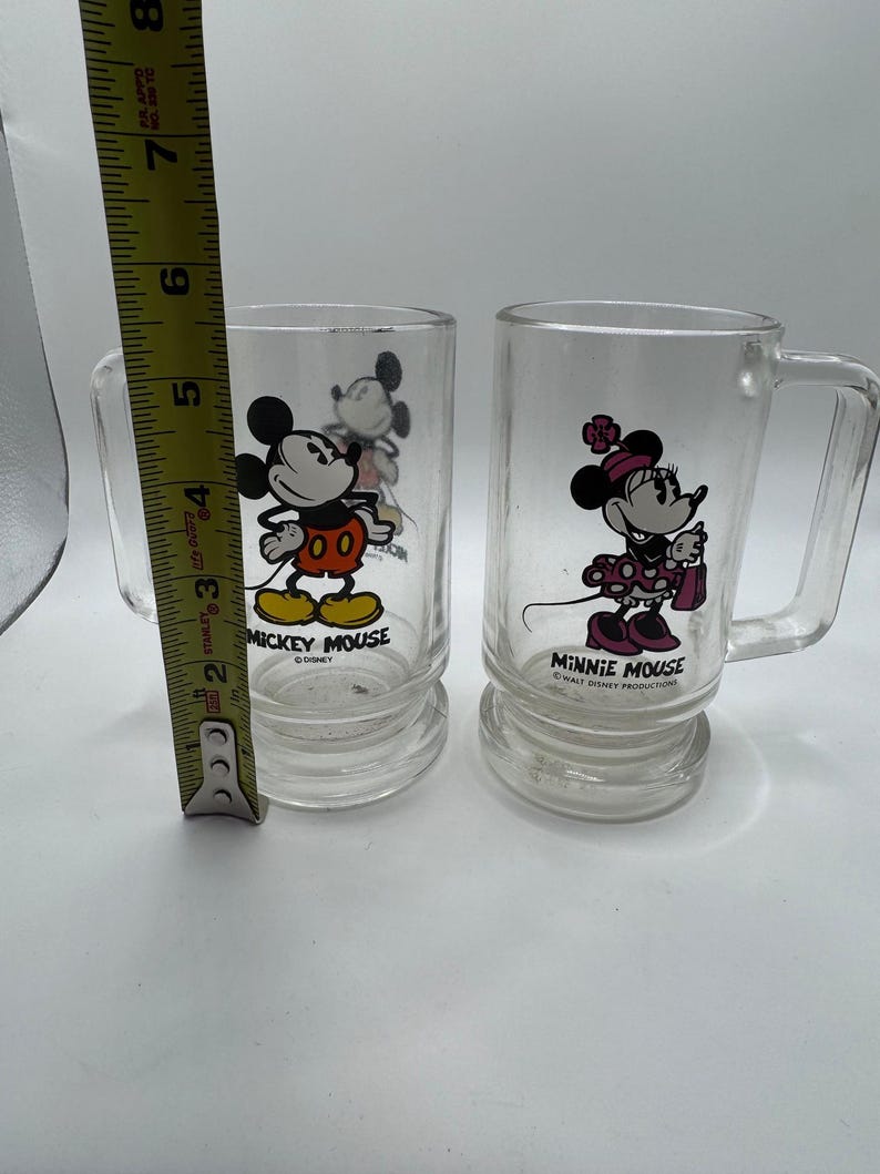 May include: Two clear glass mugs featuring Mickey Mouse and Minnie Mouse. Mickey is in his classic outfit, and Minnie is in a polka-dot dress. Each mug has a handle and the text "MICKEY MOUSE" and "MINNIE MOUSE" printed on them. The mugs are approximately 4 inches tall.