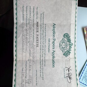 May include: A Cabbage Patch Kids Adoption Papers Application document with a green decorative border. The form includes an Oath of Adoption, a signature from the Chief of Staff, and handwritten details for Martie Fayette, dated Dec. 25, 85.