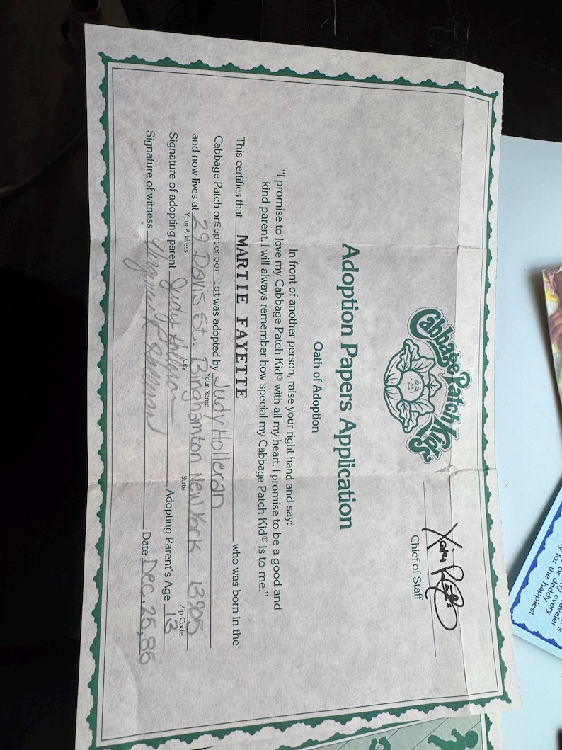May include: A Cabbage Patch Kids Adoption Papers Application document with a green decorative border. The form includes an Oath of Adoption, a signature from the Chief of Staff, and handwritten details for Martie Fayette, dated Dec. 25, 85.