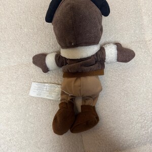 K&ouml;nnte beinhalten: Ein braunes Pl&uuml;schtier in einem Fliegeroutfit mit schwarzen Ohren. Das Spielzeug hat einen wei&szlig;en Kragen und Manschetten, braune Stiefel und eine beige Hose. Ein Etikett ist am Bein des Spielzeugs befestigt.