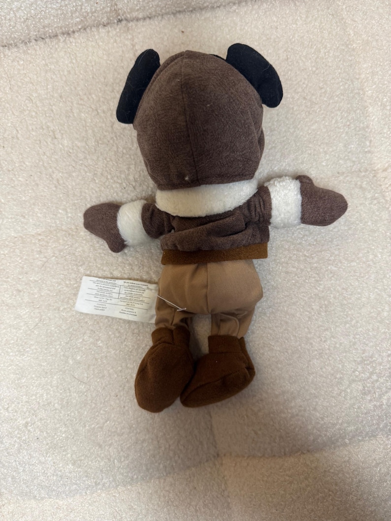 K&ouml;nnte beinhalten: Ein braunes Pl&uuml;schtier in einem Fliegeroutfit mit schwarzen Ohren. Das Spielzeug hat einen wei&szlig;en Kragen und Manschetten, braune Stiefel und eine beige Hose. Ein Etikett ist am Bein des Spielzeugs befestigt.