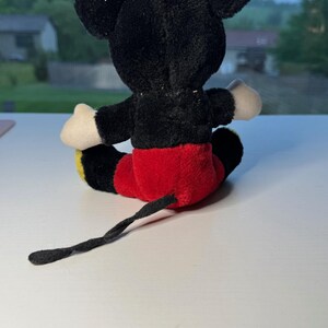 May include: A plush Mickey Mouse toy, seen from the rear. The toy has a black body, ears, and tail, with red shorts, white gloves, and yellow feet. The toy is positioned on a white surface.