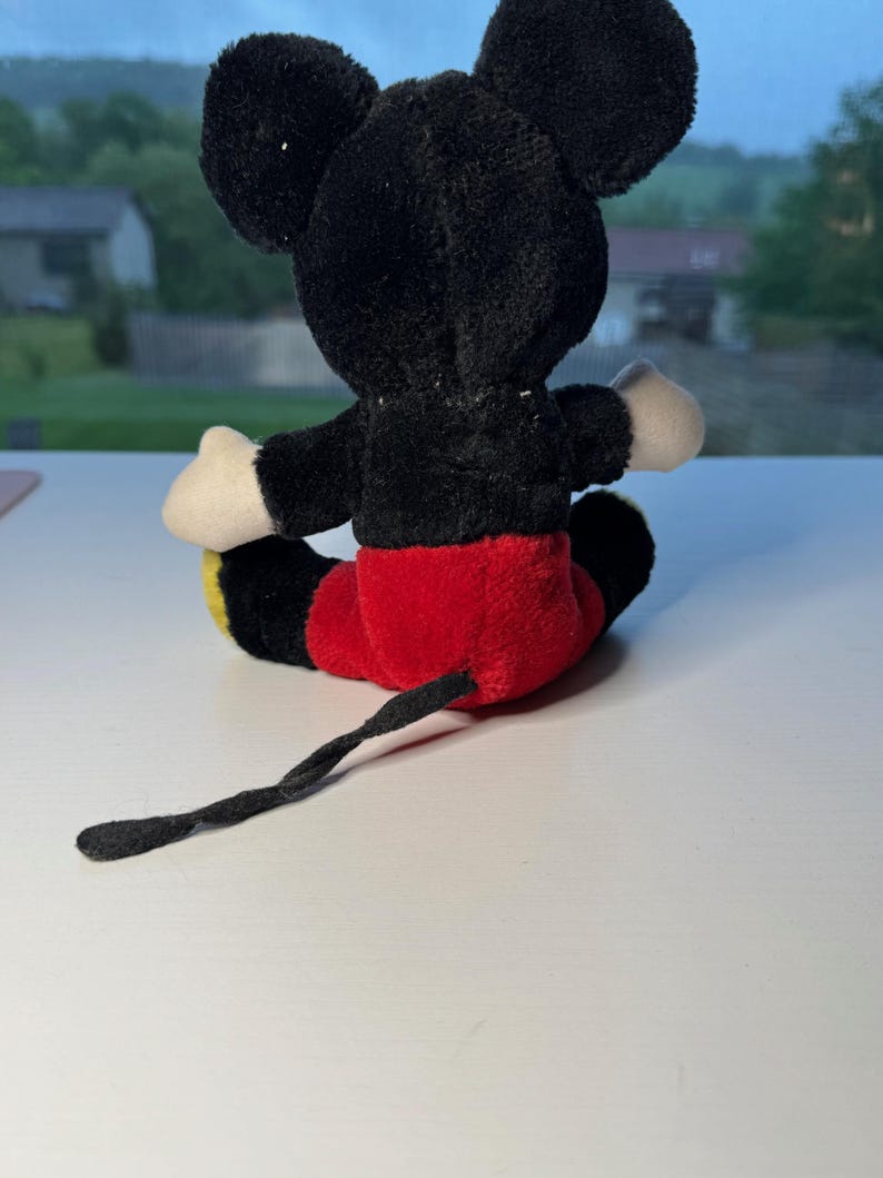 May include: A plush Mickey Mouse toy, seen from the rear. The toy has a black body, ears, and tail, with red shorts, white gloves, and yellow feet. The toy is positioned on a white surface.