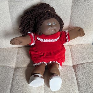 May include: A vintage doll with dark skin, brown yarn hair, and a red dress with white lace trim. The doll is wearing white shoes and is lying on a textured, off-white surface. The doll's arms are outstretched.