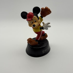 K&ouml;nnte beinhalten: Eine Mickey Mouse-Figur in Baseball-Uniform, mit roter Kappe, gelbem Hemd und roter Hose. Er posiert, als w&uuml;rde er einen Ball mit einem braunen Handschuh fangen. Die Figur steht auf einem schwarzen Sockel mit dem Wort "SUPER".