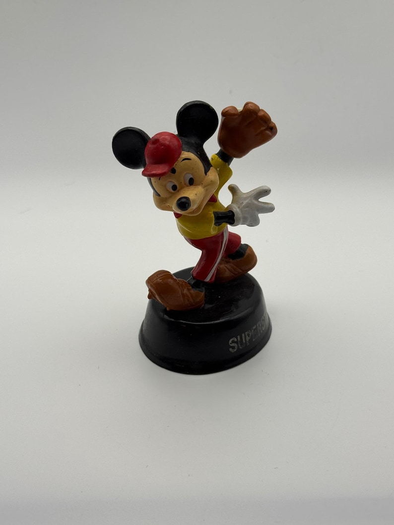 K&ouml;nnte beinhalten: Eine Mickey Mouse-Figur in Baseball-Uniform, mit roter Kappe, gelbem Hemd und roter Hose. Er posiert, als w&uuml;rde er einen Ball mit einem braunen Handschuh fangen. Die Figur steht auf einem schwarzen Sockel mit dem Wort "SUPER".