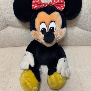 Puede incluir: Mu&ntilde;eca de peluche de Minnie Mouse con pelaje negro, orejas negras grandes y un lazo rojo con lunares blancos. La mu&ntilde;eca tiene una cara bronceada, guantes blancos y pies amarillos. La mu&ntilde;eca est&aacute; sentada.