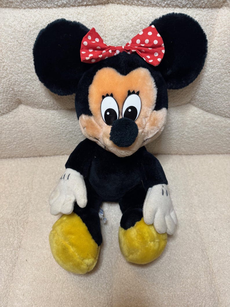 Puede incluir: Mu&ntilde;eca de peluche de Minnie Mouse con pelaje negro, orejas negras grandes y un lazo rojo con lunares blancos. La mu&ntilde;eca tiene una cara bronceada, guantes blancos y pies amarillos. La mu&ntilde;eca est&aacute; sentada.