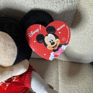 May include: A Mickey Mouse plush toy with a red heart-shaped tag featuring Mickey's face. The plush has black ears, a red outfit, and white gloves. The tag reads "Disney" and "Mickey".