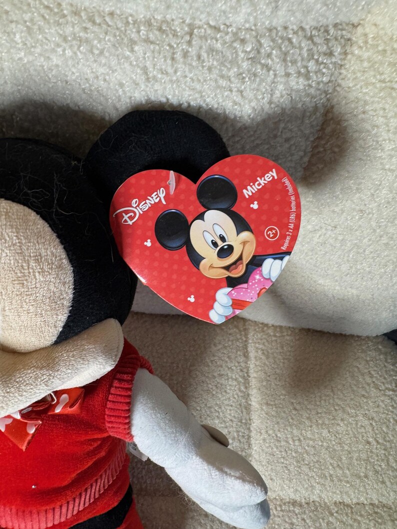 May include: A Mickey Mouse plush toy with a red heart-shaped tag featuring Mickey's face. The plush has black ears, a red outfit, and white gloves. The tag reads "Disney" and "Mickey".