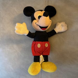 May include: A black and red plush Mickey Mouse toy with yellow shoes. The toy is standing on a gray surface.