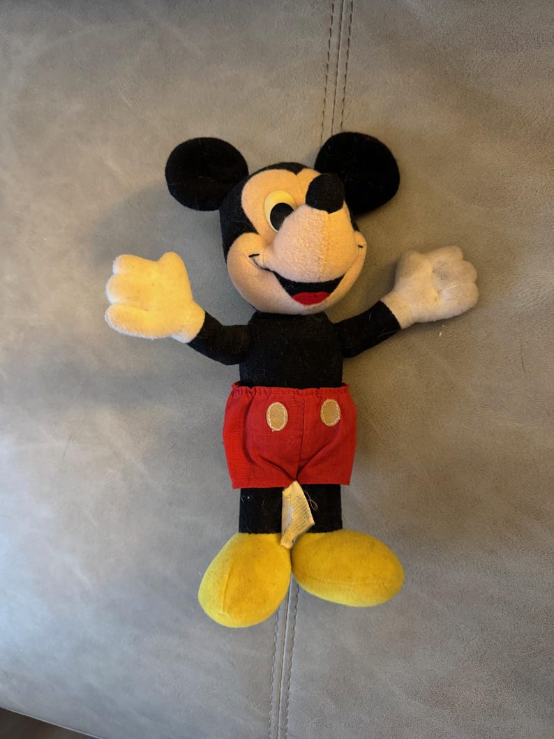 May include: A black and red plush Mickey Mouse toy with yellow shoes. The toy is standing on a gray surface.