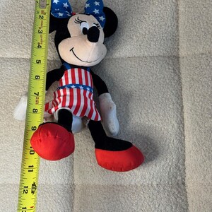 May include: A Minnie Mouse soft toy, approximately 33 cm tall, with a patriotic theme. The toy features a red, white, and blue colour scheme, including a bow with white stars on a blue background, and a dress with red and white stripes.