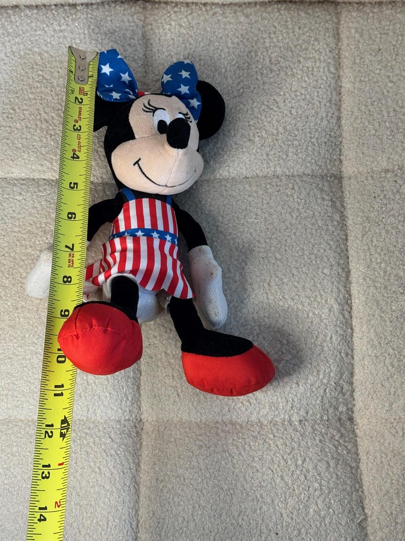 May include: A Minnie Mouse soft toy, approximately 33 cm tall, with a patriotic theme. The toy features a red, white, and blue colour scheme, including a bow with white stars on a blue background, and a dress with red and white stripes.
