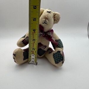 May include: A small, beige teddy bear with patchwork accents on its paws and ears. The bear has a burgundy plaid bow and measures approximately 6 inches tall.