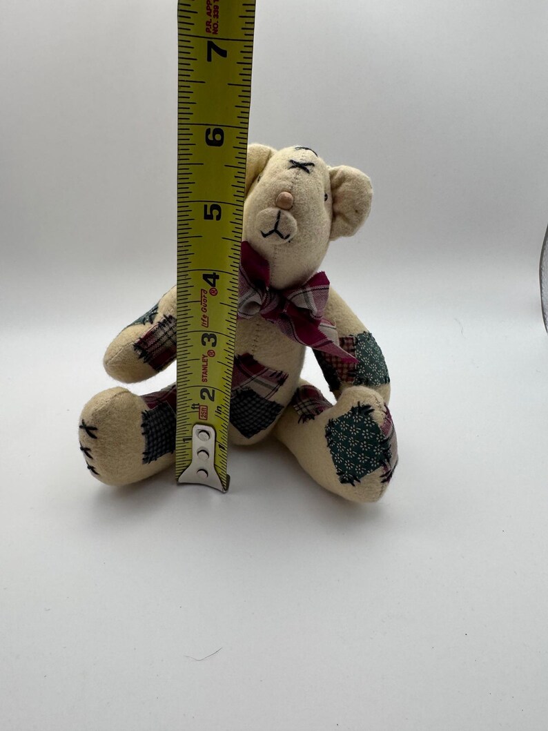 May include: A small, beige teddy bear with patchwork accents on its paws and ears. The bear has a burgundy plaid bow and measures approximately 6 inches tall.