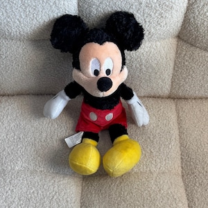 May include: A plush Mickey Mouse toy. The toy has black ears, a black head, and a tan face. It is wearing red shorts, yellow shoes, and white gloves. The toy is sitting on a cream-colored, textured surface.