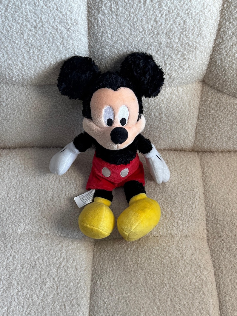 May include: A plush Mickey Mouse toy. The toy has black ears, a black head, and a tan face. It is wearing red shorts, yellow shoes, and white gloves. The toy is sitting on a cream-colored, textured surface.
