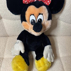 Puede incluir: Mu&ntilde;eca de peluche de Minnie Mouse con pelaje negro, grandes orejas negras y un lazo rojo con lunares blancos. La mu&ntilde;eca tiene una cara bronceada, guantes blancos y pies amarillos. Un juguete infantil cl&aacute;sico.