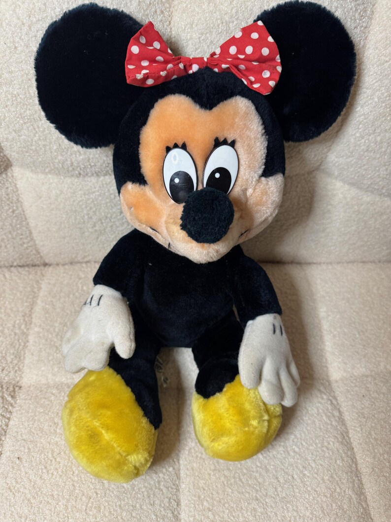 Puede incluir: Mu&ntilde;eca de peluche de Minnie Mouse con pelaje negro, grandes orejas negras y un lazo rojo con lunares blancos. La mu&ntilde;eca tiene una cara bronceada, guantes blancos y pies amarillos. Un juguete infantil cl&aacute;sico.