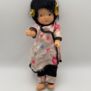 May include: A vintage doll with black hair and yellow flower accents. The doll wears a pink floral patterned top with black trim and black pants. The doll's outfit has pink floral accents. The doll's skin is a light peach color.