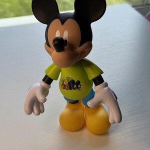 May include: A Mickey Mouse figurine with black ears, a tan face, and a black nose. Mickey wears a green shirt with a graphic, blue shorts, yellow shoes, and white gloves. The figurine is standing on a white surface.