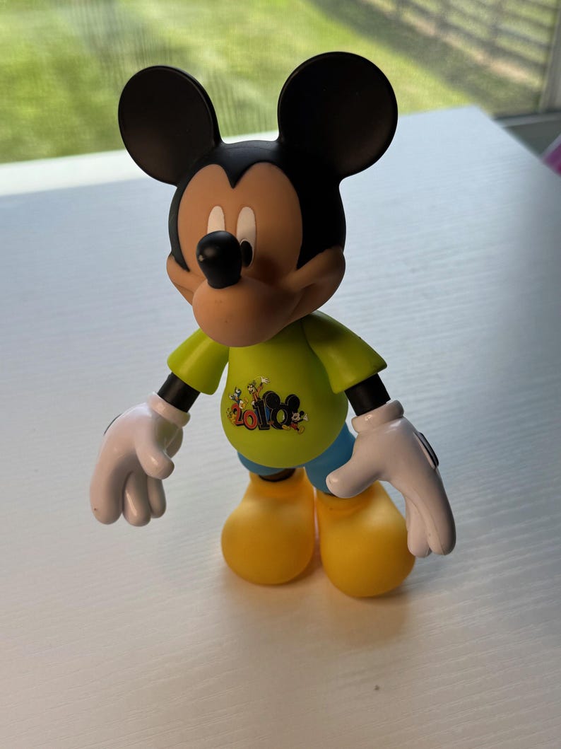 May include: A Mickey Mouse figurine with black ears, a tan face, and a black nose. Mickey wears a green shirt with a graphic, blue shorts, yellow shoes, and white gloves. The figurine is standing on a white surface.