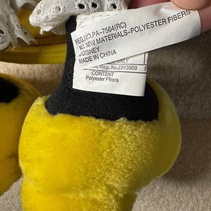 May include: Close-up of a plush toy's foot, featuring bright yellow fur and a black base. A white tag with text, including "DISNEY" and "Polyester Fibers," is visible. The toy's design is partially visible, with white fabric and lace.