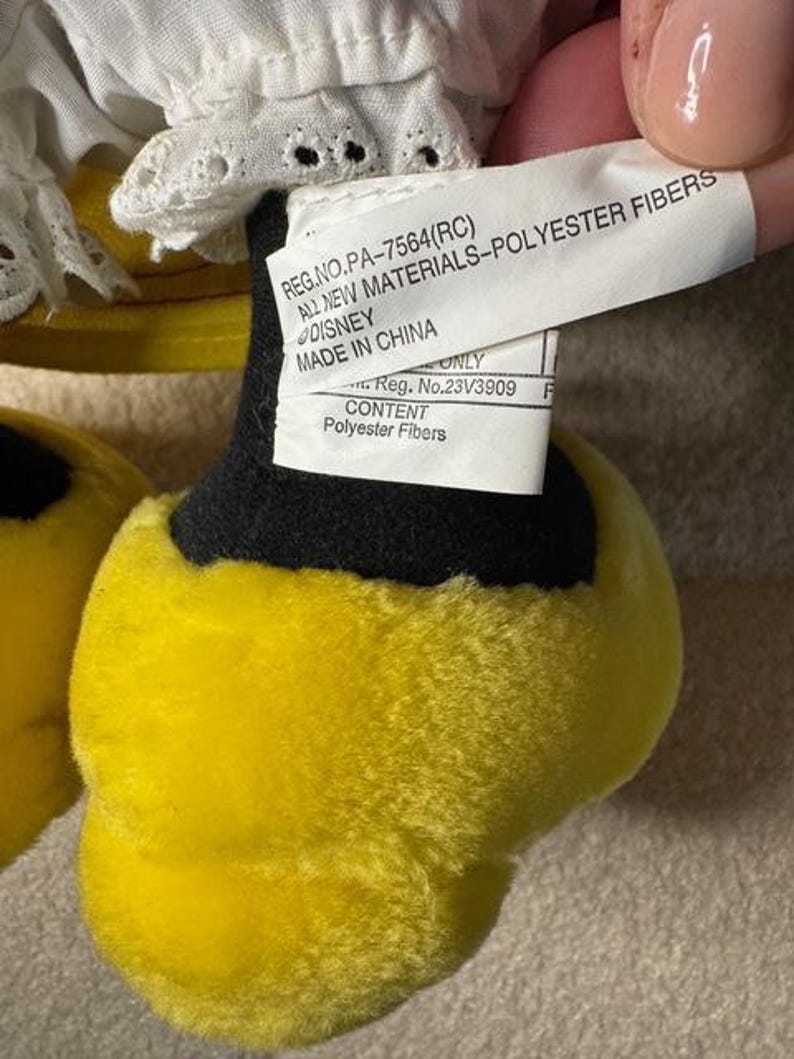 May include: Close-up of a plush toy's foot, featuring bright yellow fur and a black base. A white tag with text, including "DISNEY" and "Polyester Fibers," is visible. The toy's design is partially visible, with white fabric and lace.