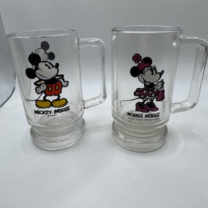 May include: Two clear glass mugs featuring Mickey Mouse and Minnie Mouse. Mickey is in his classic outfit, and Minnie is in a polka-dot dress. Each mug has a handle and a base with the character's name printed below.