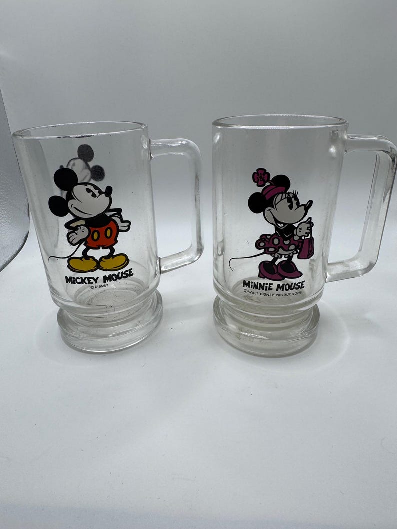 May include: Two clear glass mugs featuring Mickey Mouse and Minnie Mouse. Mickey is in his classic outfit, and Minnie is in a polka-dot dress. Each mug has a handle and a base with the character's name printed below.