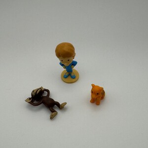 May include: Three small figurines: a boy in blue, a brown monkey, and an orange tiger. The boy stands on a yellow base. The monkey is lying down. The tiger has black stripes. These are likely collectible toys.