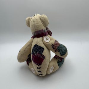 May include: A beige teddy bear with patchwork accents on its arms and legs. The bear has a plaid scarf around its neck and button details. The bear is a handmade item.