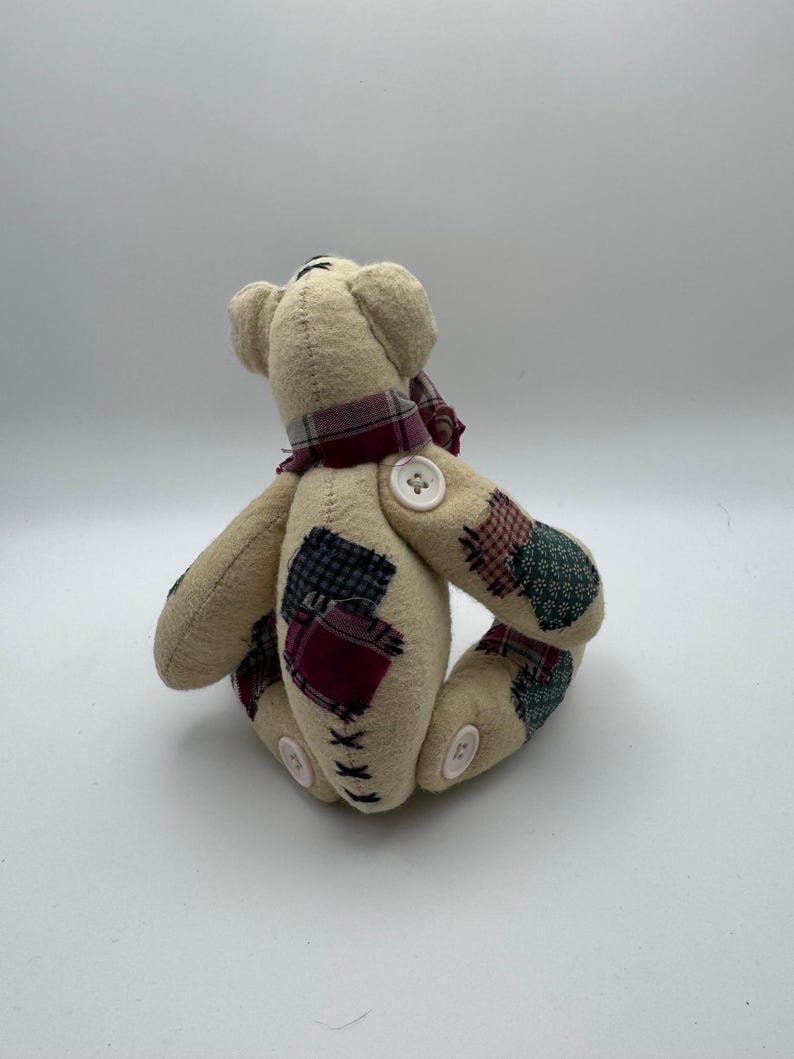 May include: A beige teddy bear with patchwork accents on its arms and legs. The bear has a plaid scarf around its neck and button details. The bear is a handmade item.