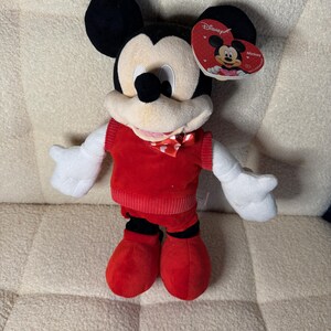 May include: A plush Mickey Mouse toy. The toy is wearing a red sweater, shorts, and shoes. Mickey has white gloves, a cream-colored face, and black ears. A heart-shaped tag with the Disney logo is attached.