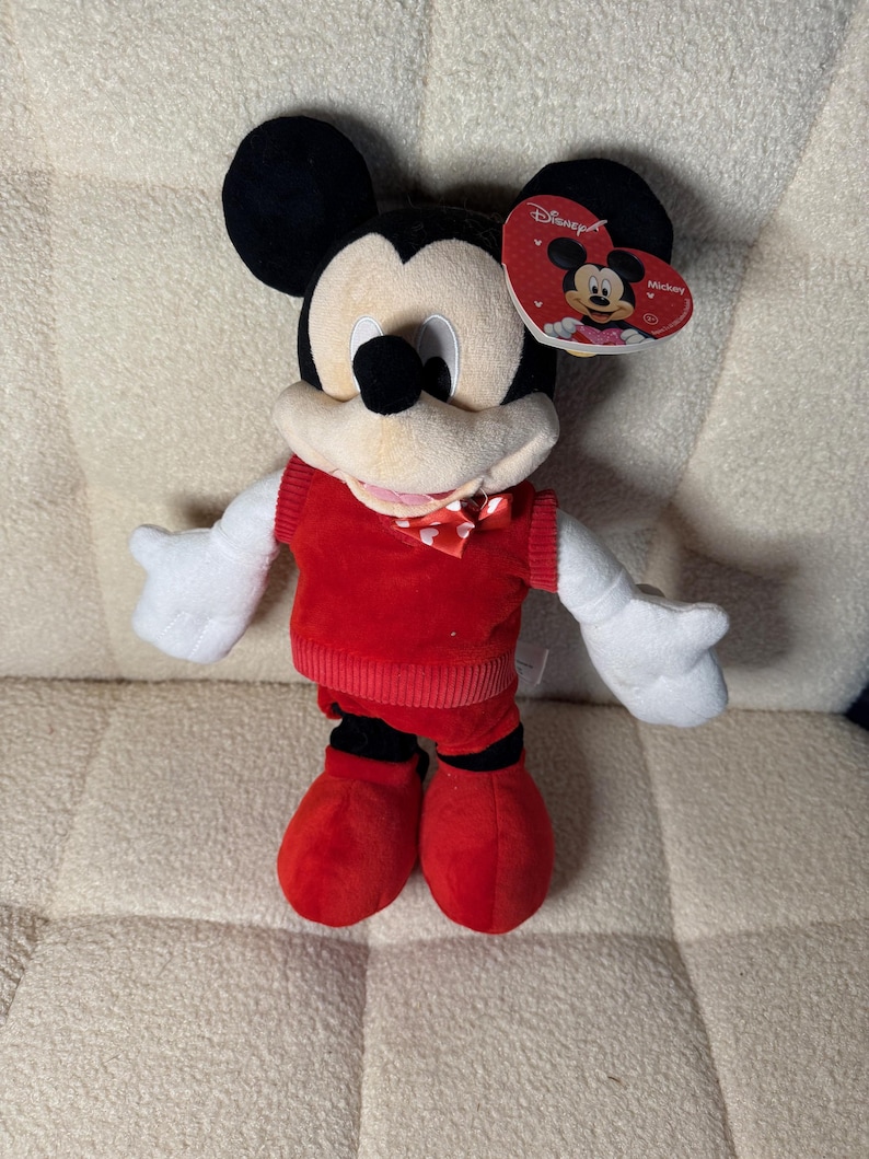 May include: A plush Mickey Mouse toy. The toy is wearing a red sweater, shorts, and shoes. Mickey has white gloves, a cream-colored face, and black ears. A heart-shaped tag with the Disney logo is attached.